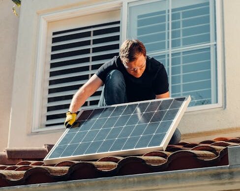A worker installing a solar panel on a residential rooftop under bright sunlight.